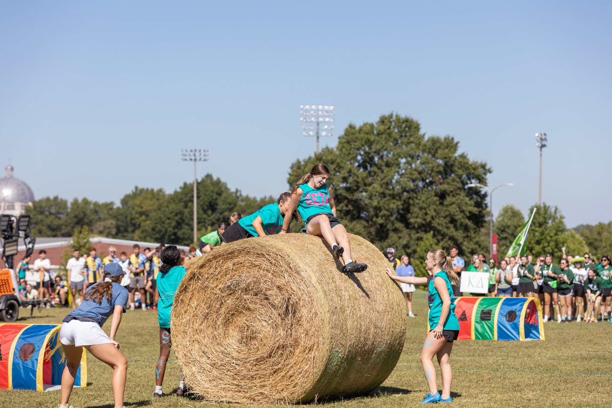 Several young people in teal shirts compete in an outdoor event, climbing over a large round hay bale on a grassy field, with spectators and colorful obstacles in the background under a clear sky.
