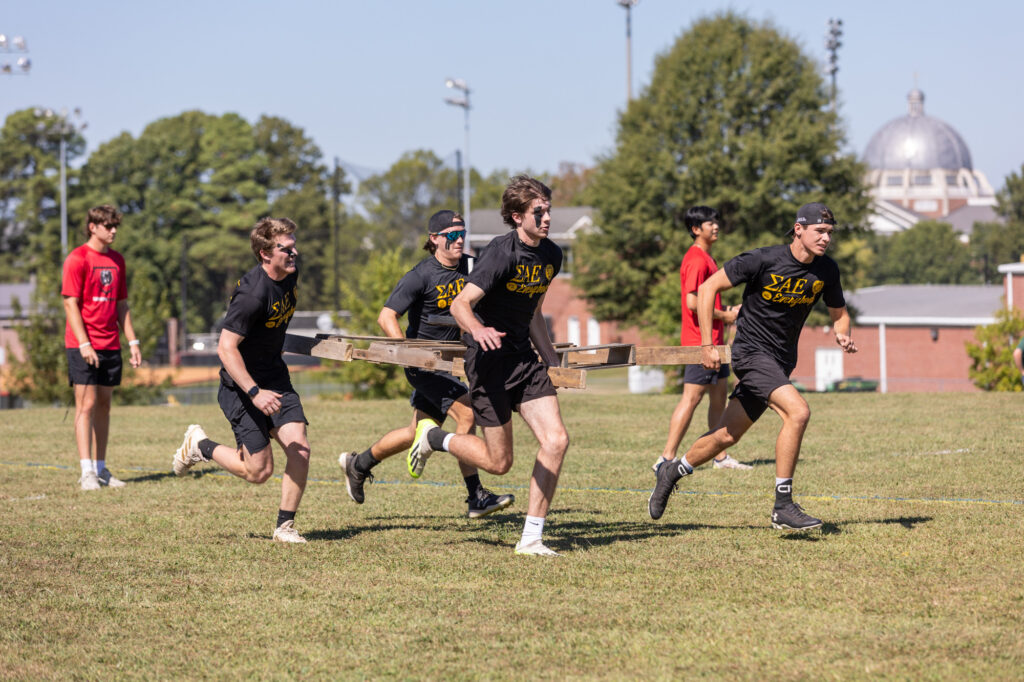 Six young men run across a grassy field, five of them carrying wooden pallets. Most wear black T-shirts with Greek letters, while one in the background wears red. There are trees and a building with a dome visible in the background.