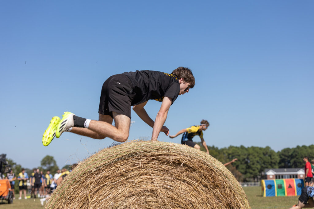 A person in athletic clothes climbs over a large round hay bale during an outdoor obstacle race, with other participants and spectators visible in the background under a clear sky.