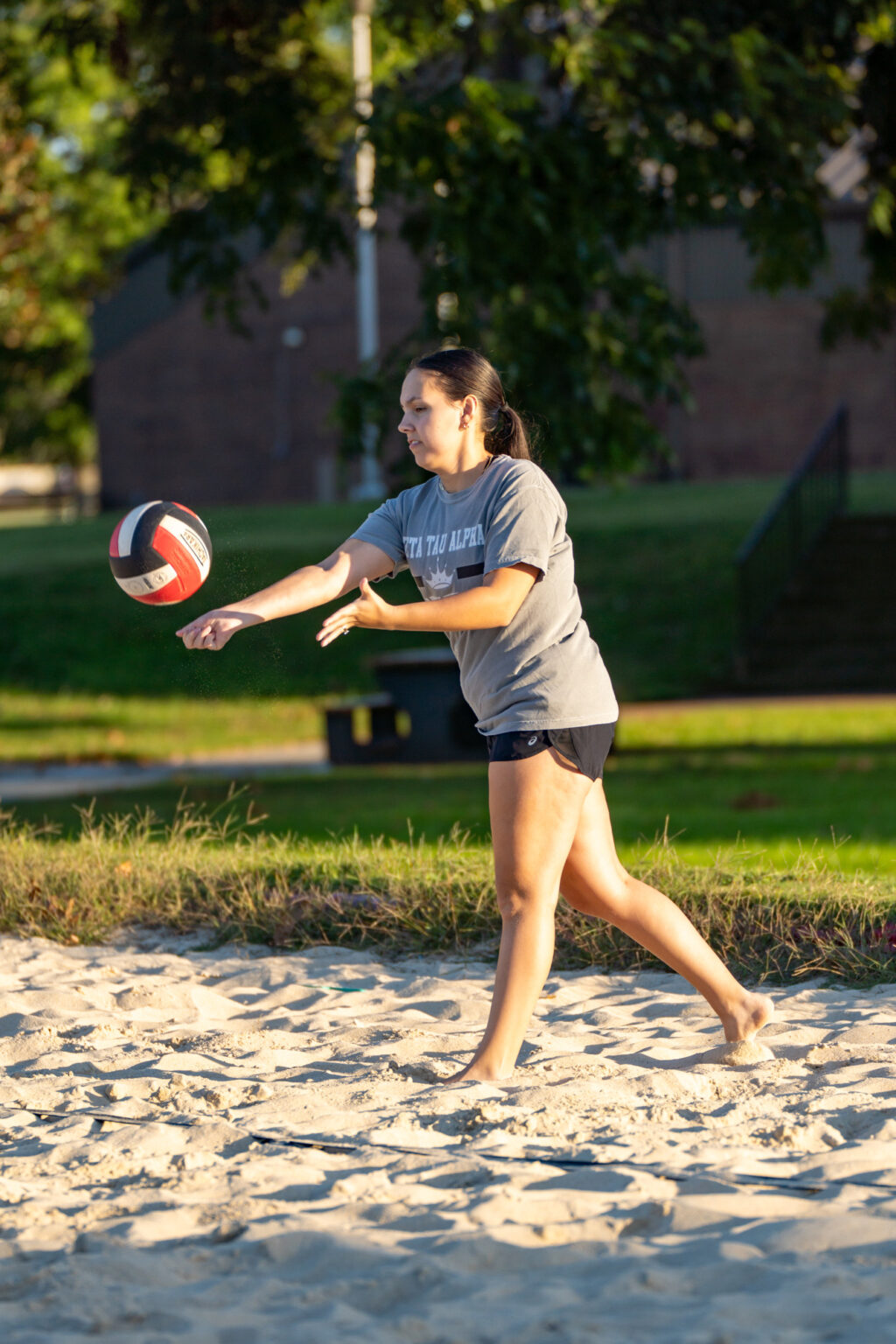 A woman in a gray T-shirt and black shorts stands barefoot on a sandy court, serving a volleyball with her right hand. There is grass and a building in the background.