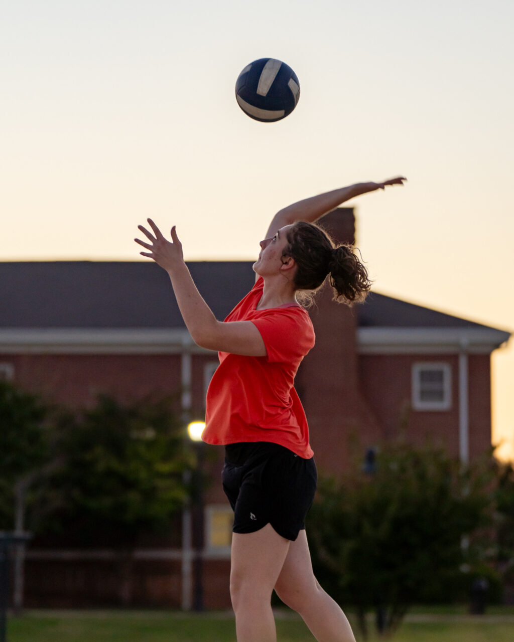 A person in a red shirt and black shorts is serving a volleyball outdoors, with one arm raised and the ball in mid-air. A building and trees are visible in the background at dusk.