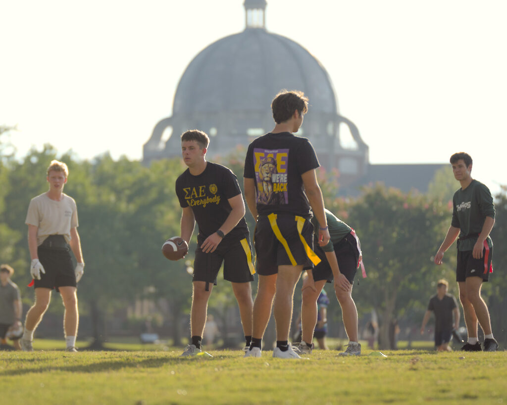 A group of young men play flag football on a grassy field. One holds a football, preparing to snap. They wear casual athletic clothes, some with colored flags. A large domed building is visible in the background.