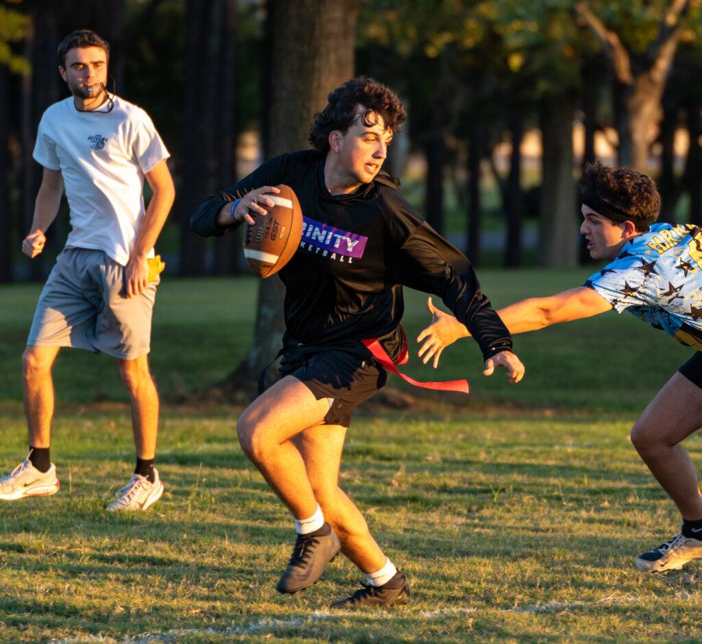Three people play flag football on grass. One player in a black shirt holds a football and tries to evade a defender reaching for his flag. Another player in a white shirt watches from behind. Trees are visible in the background.