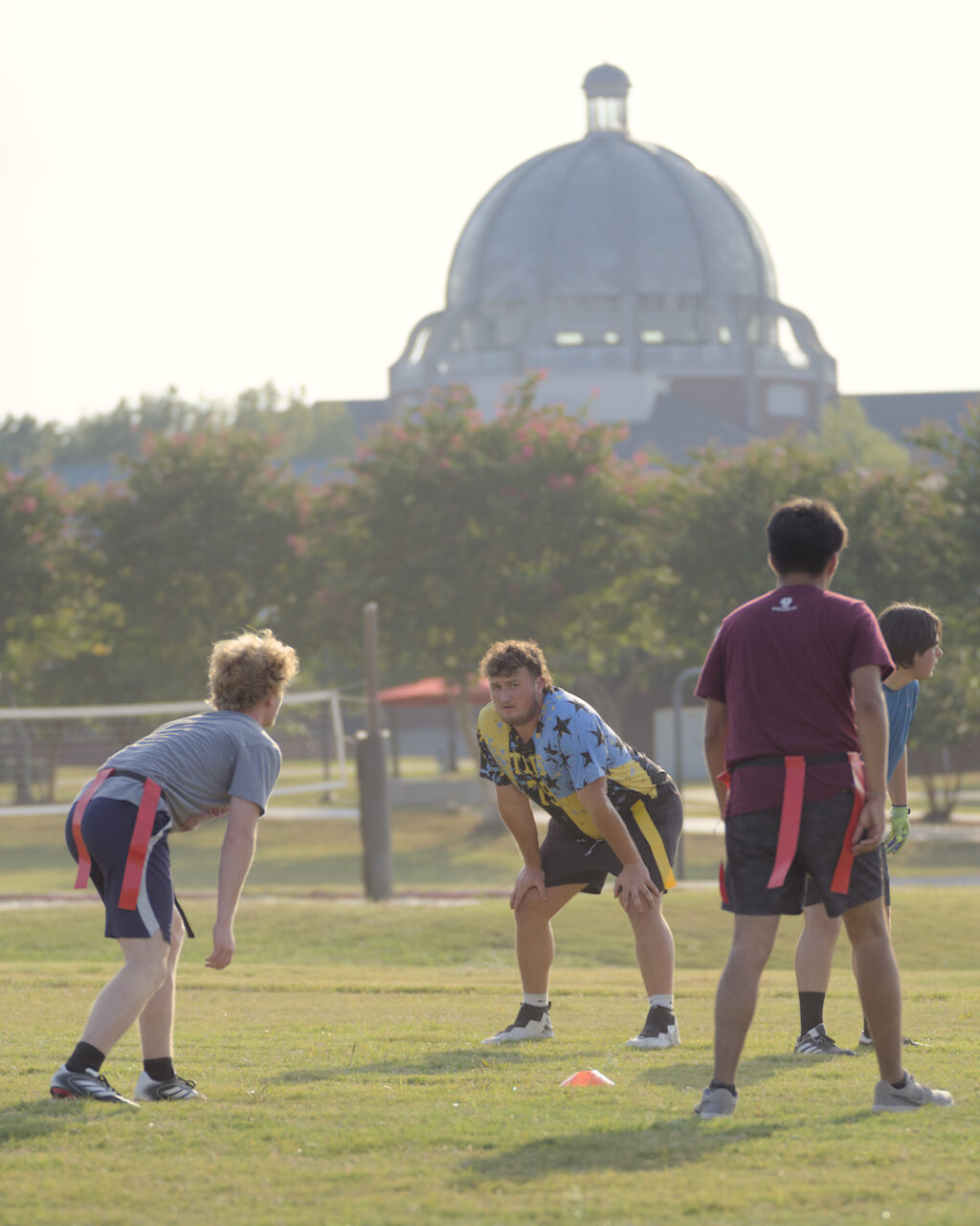 Four people play flag football on a grassy field. Two are facing each other, ready for play, while two stand nearby. A domed building and trees are visible in the background. The weather appears sunny and warm.
