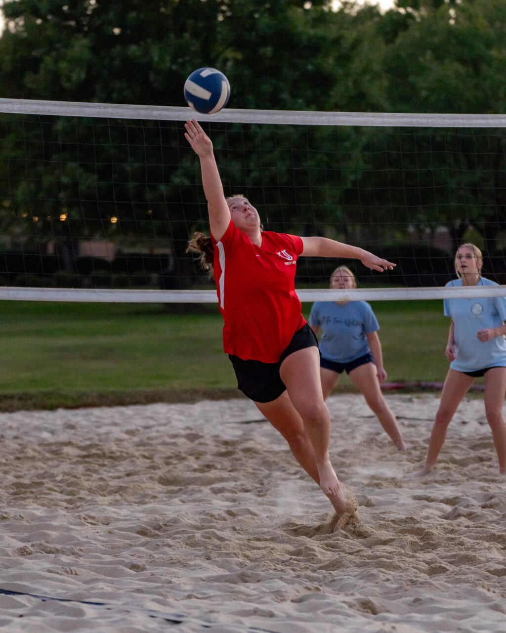 A person in a red shirt jumps on a sand volleyball court, reaching with one arm toward a volleyball above the net. Two other players in blue shirts watch from behind. Trees and grass are visible in the background.