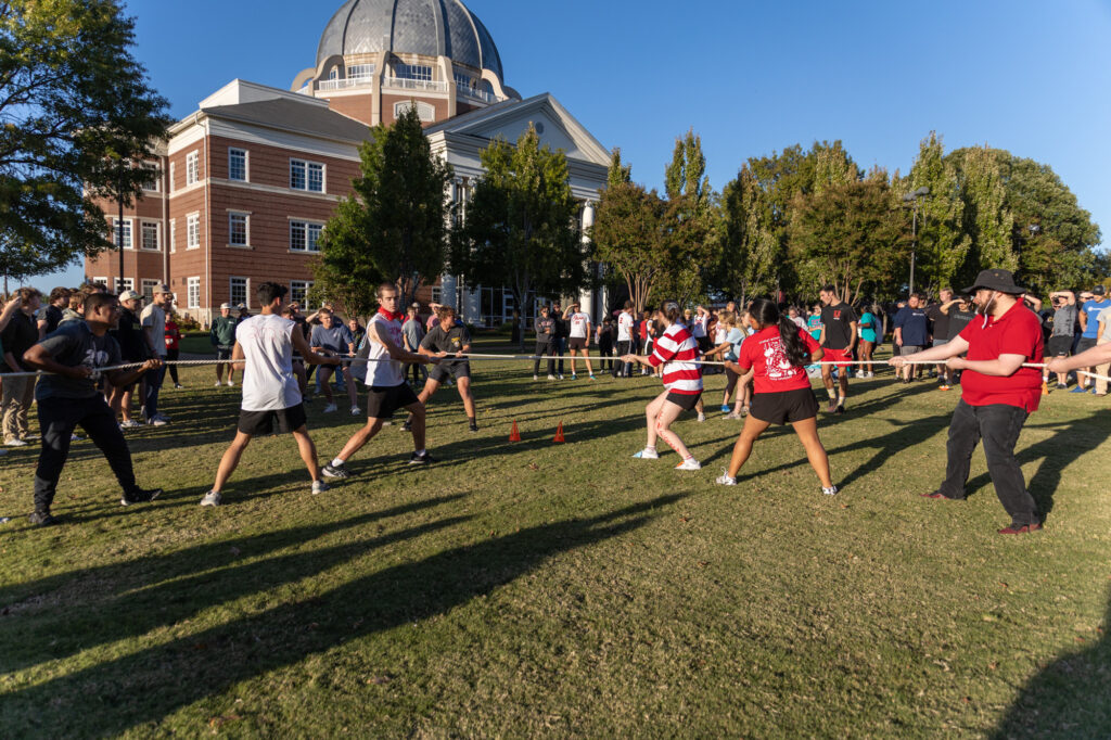 People participate in a tug-of-war game on a grassy field with a crowd watching. A large, domed building and trees are in the background on a sunny day.