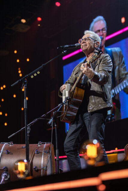 An older man wearing glasses and a brown jacket plays an acoustic guitar and sings into a microphone on stage, with stage lights and a large drum in the background.