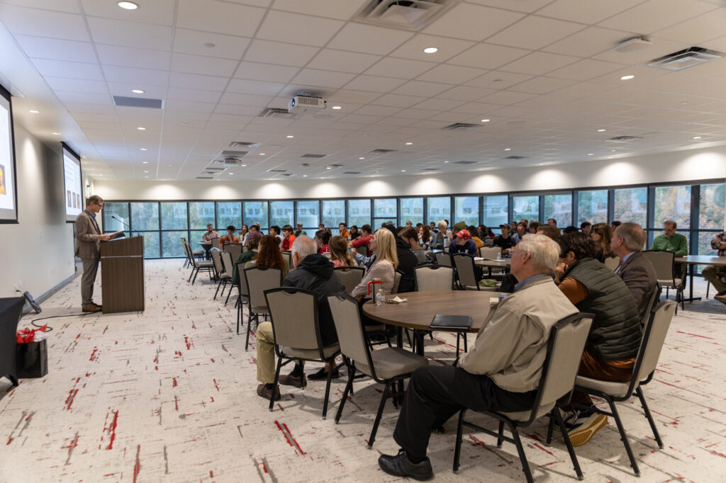 A speaker stands at a podium addressing a seated audience in a large, well-lit conference room with windows lining one wall. Attendees face the speaker, listening attentively.