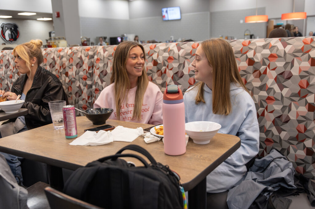 Two young women sit at a table in a cafeteria, talking and smiling. There are bowls, a tall pink water bottle, a drink can, and napkins on the table. Another person sits nearby, eating. The background shows cafeteria counters.