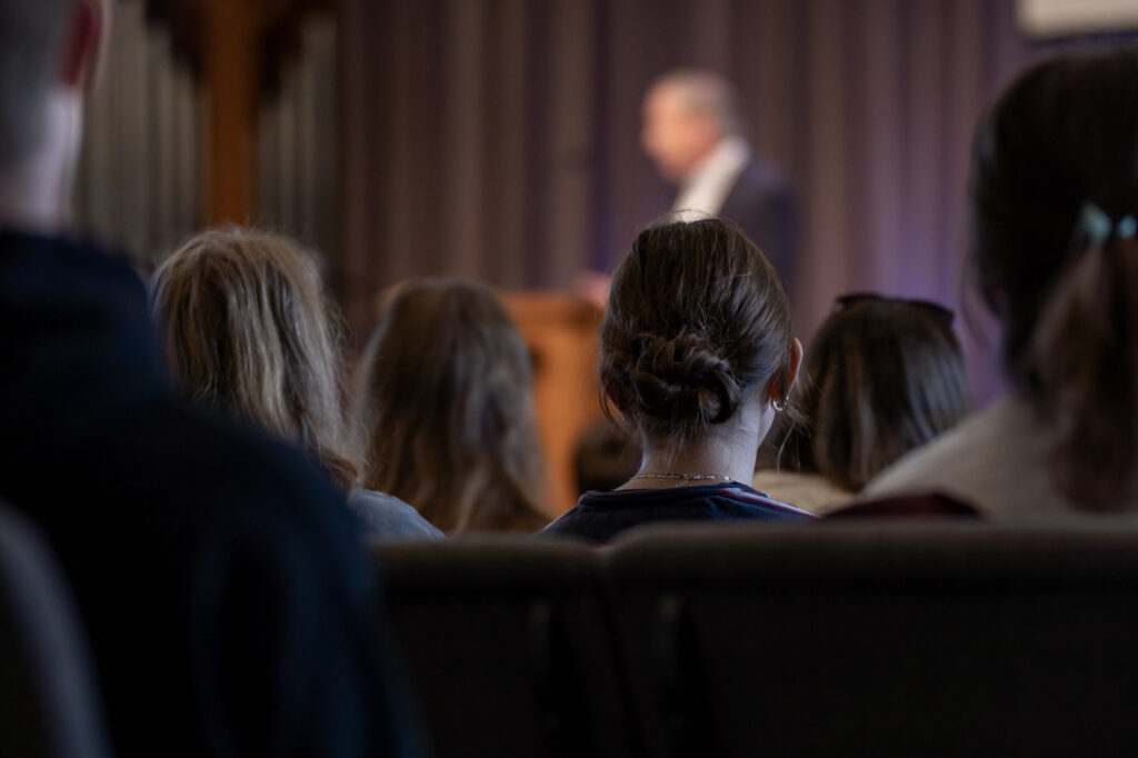 A group of people sit and face a blurred speaker at the front of a room with dark curtains. The focus is on the audience, viewed from behind, while the speaker remains out of focus in the background.