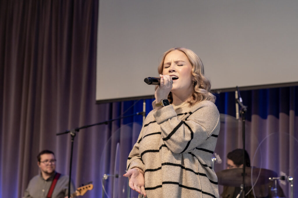 A woman sings into a microphone on stage, wearing a striped sweater. A band with a guitarist and drummer plays behind her. The background includes curtains and a large screen.