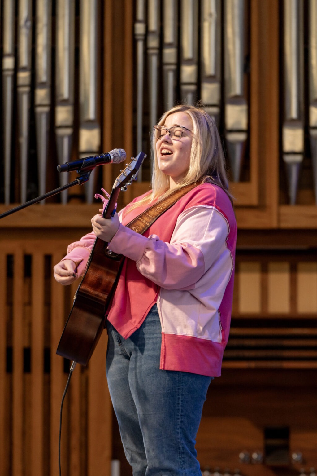 A person wearing glasses, a pink jacket, and jeans plays an acoustic guitar and sings into a microphone onstage. Large organ pipes are visible in the background.