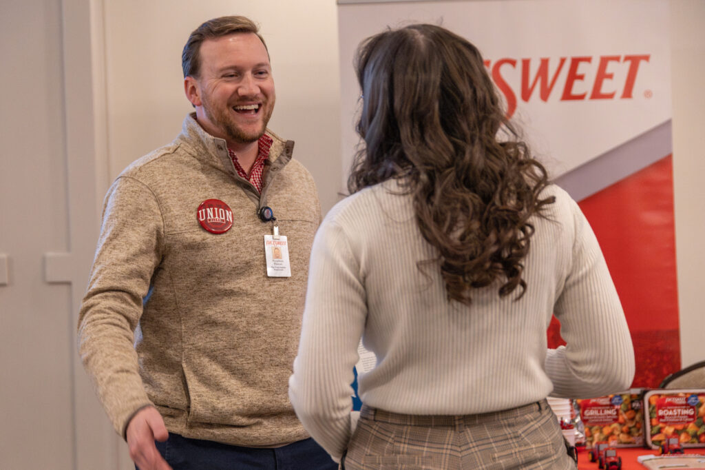 A man wearing a Union sticker and a lanyard speaks and smiles at a woman in a plaid skirt. In the background, a display table and a banner reading SWEET are visible, suggesting an event organized by the Vocatio Center for Life Calling and Career.