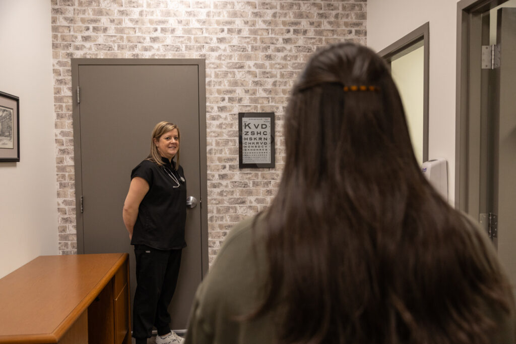 A medical professional stands by a door in an exam room, facing a patient with long brown hair. An eye chart is mounted on the brick-patterned wall between them.