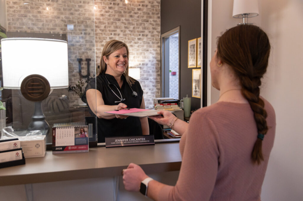 A woman wearing a stethoscope hands a pink paper to another woman across a reception counter with a nameplate that reads “Jennifer Chikazte.” The scene takes place in an office with brick walls and framed pictures.