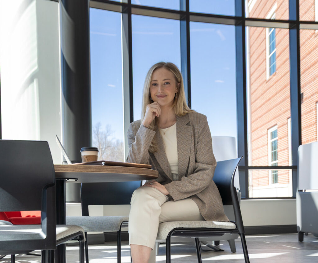 A woman sits at a round table in a modern, sunlit room with large windows. She is wearing a beige suit and white sneakers, with a coffee cup and laptop on the table, and is smiling at the camera.