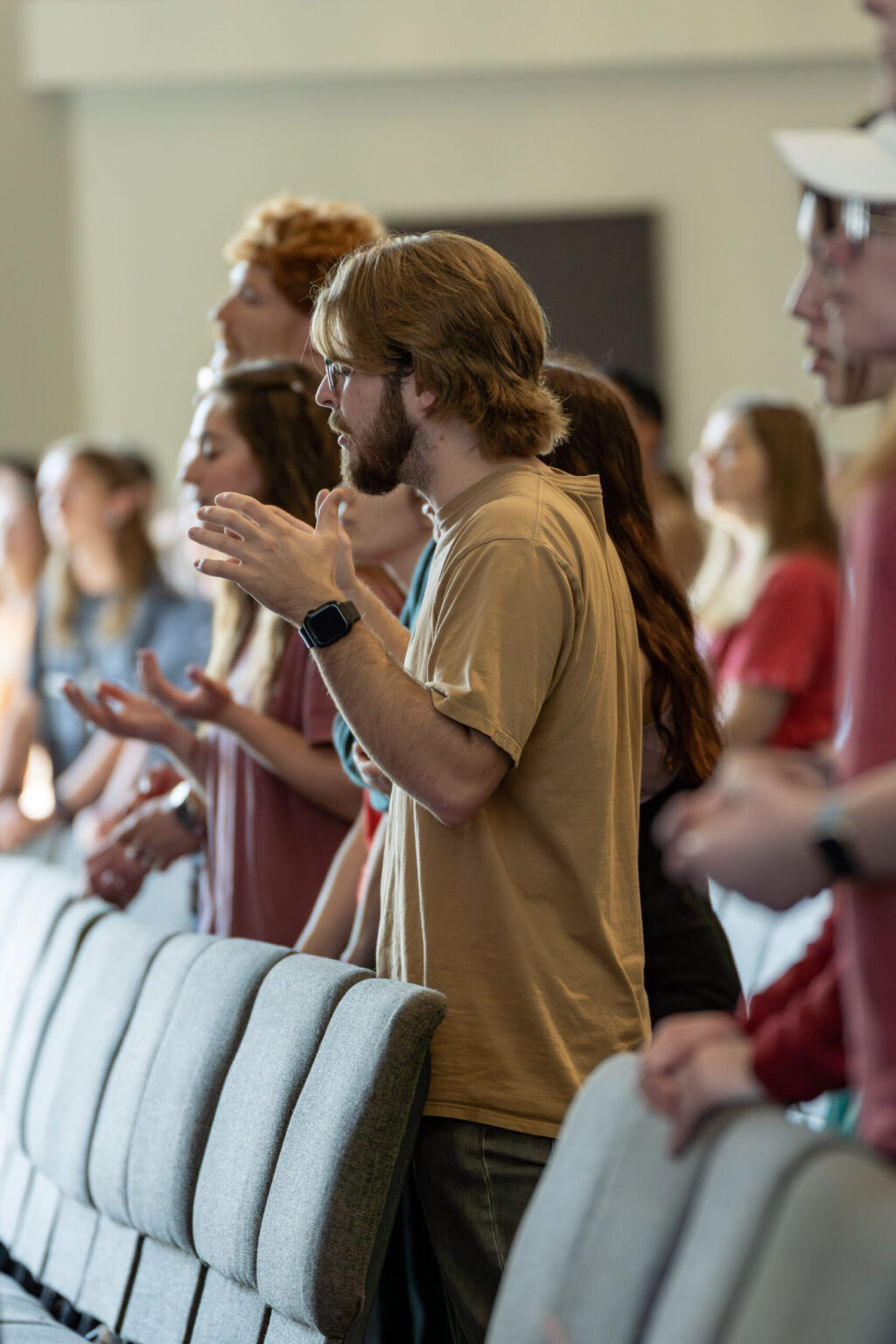 A group of people stand in rows of seats, facing forward. A man in the foreground wears glasses and a smart watch, with his hands raised. Others nearby also have their hands raised. The setting appears to be indoors.