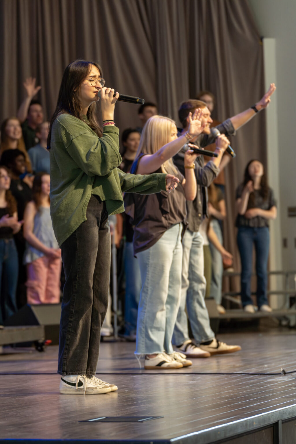 Several young adults stand on stage holding microphones and singing, with people in the background clapping and singing along. The performers are casually dressed, and the stage has a brown curtain as a backdrop.