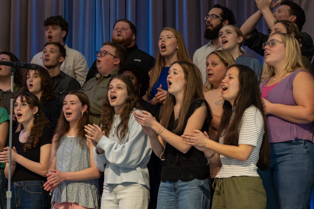 A group of people stand closely together on stage, singing with their mouths open and hands clapping. They appear focused and engaged, with a microphone visible in front of them. A blue curtain hangs in the background.