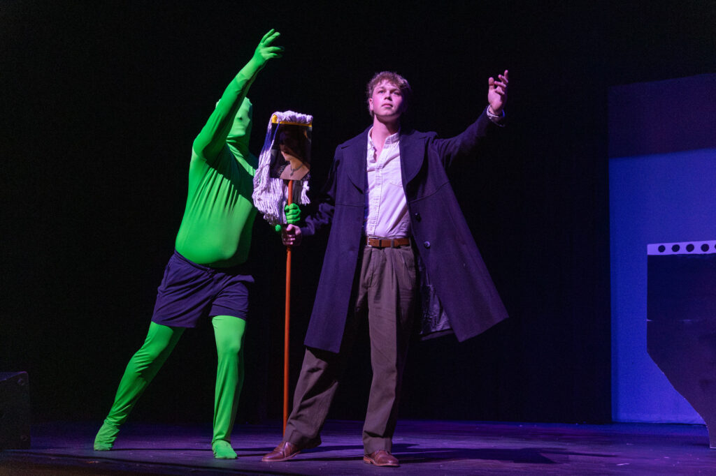 Two performers are on stage. One wears a full green bodysuit and holds a mop, while the other wears a dark coat, shirt, and trousers, gesturing with one hand. The background is dark with minimal set pieces.