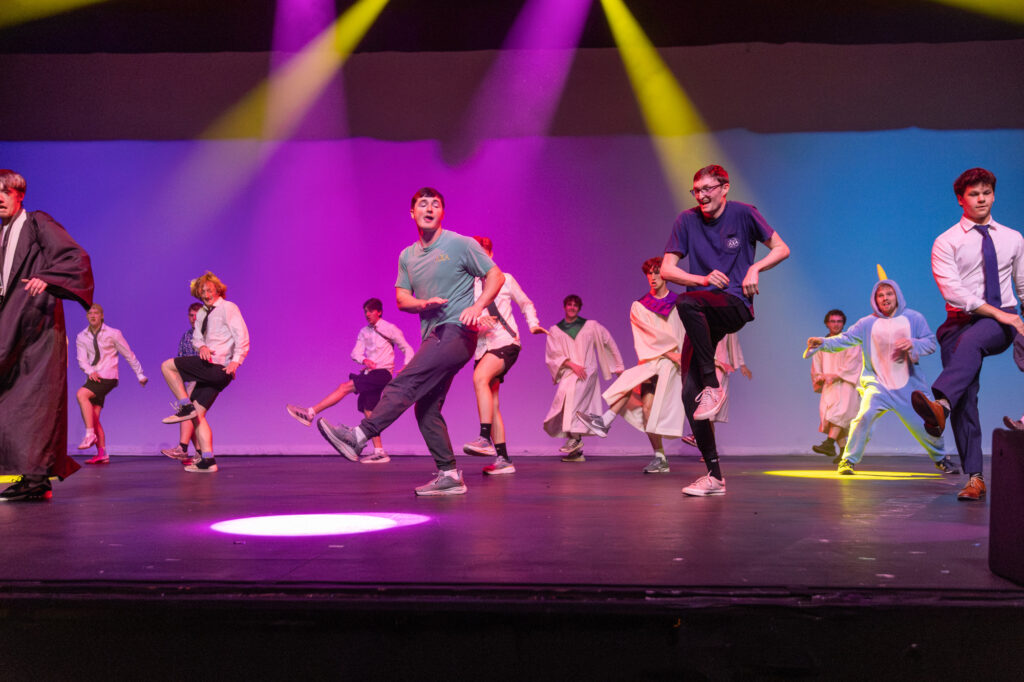 A group of people perform a choreographed dance on stage under colorful lights, wearing casual clothes, costumes, and uniforms, with some kicking one leg up in unison.