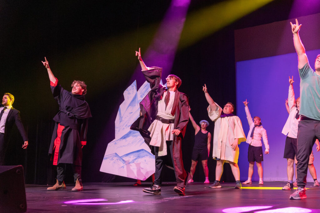 A group of people on stage perform with one arm raised, wearing a mix of costumes and casual clothing. Stage lighting casts colored beams, and abstract set pieces are visible in the background.