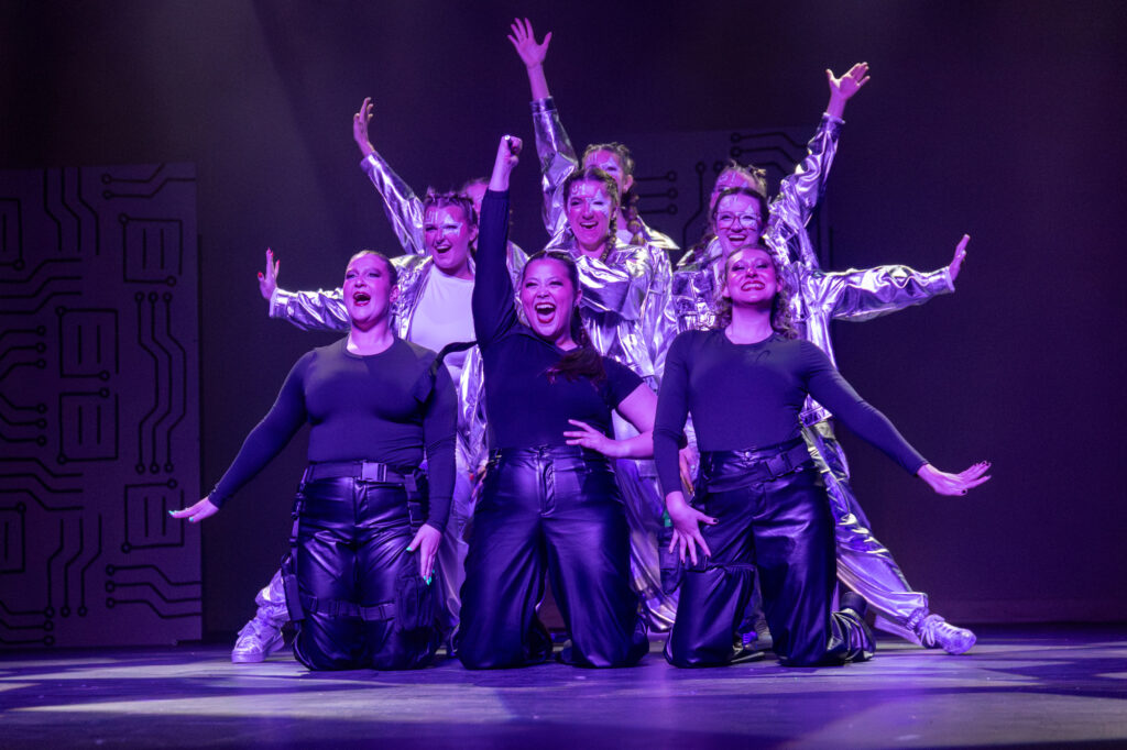 Eight performers are on stage under purple lighting. The front row wears black tops and shiny black pants, while the back row wears shiny silver outfits. They pose energetically with arms raised and smiling expressions.