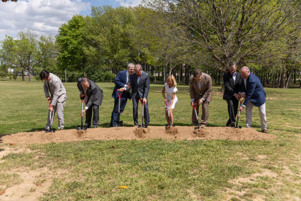 Eight people in formal attire, including one girl in a white dress, stand in a row outdoors, each holding a shovel and breaking ground on a dirt strip in a grassy area with trees in the background.