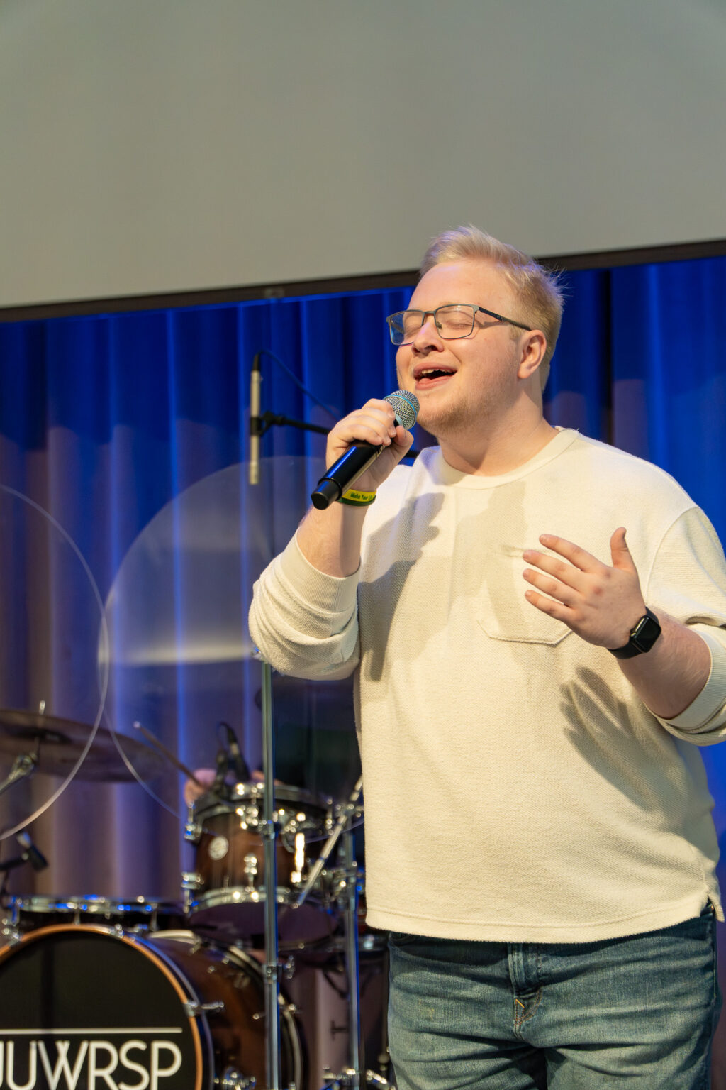 A person wearing glasses and a white shirt sings into a microphone on stage. A drum set is visible in the background. The person appears focused, with one hand raised and eyes closed. Blue curtains hang behind the stage.