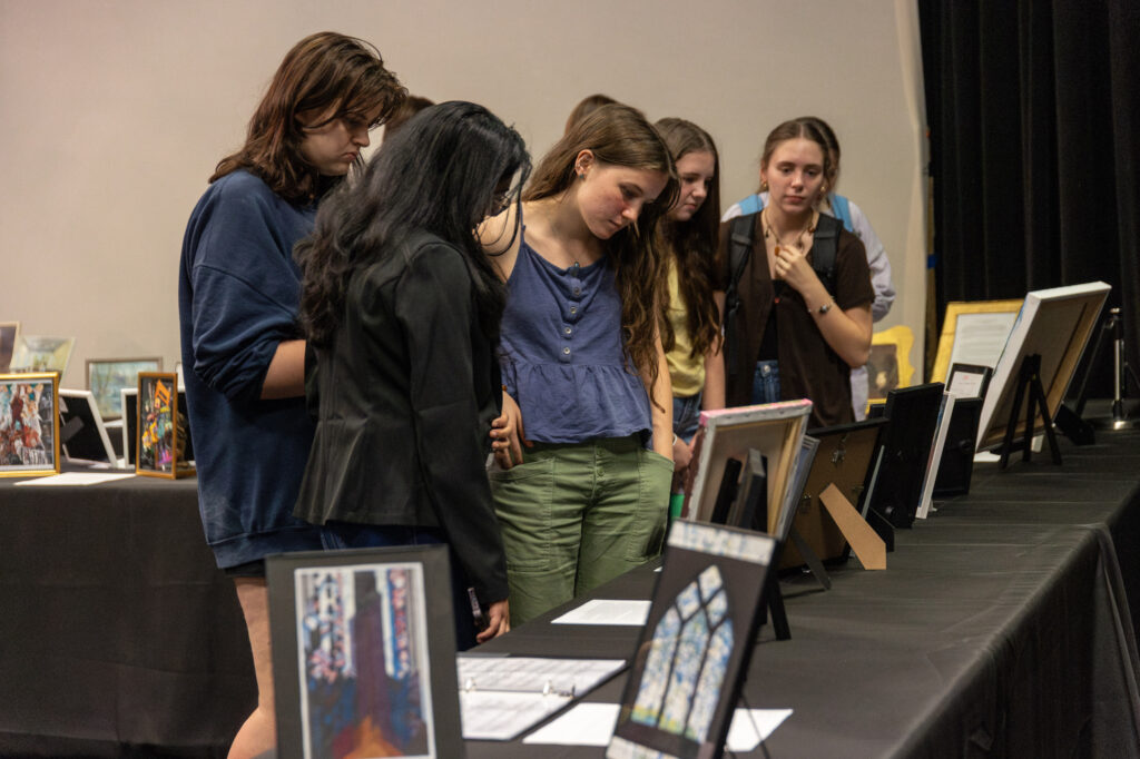Five people stand closely together, looking at framed pictures and documents displayed on easels and tables in an indoor exhibition setting. The focused figures appear to be members of the Honors Community, engaged in thoughtful exploration of the displays in this neutral, inviting environment.