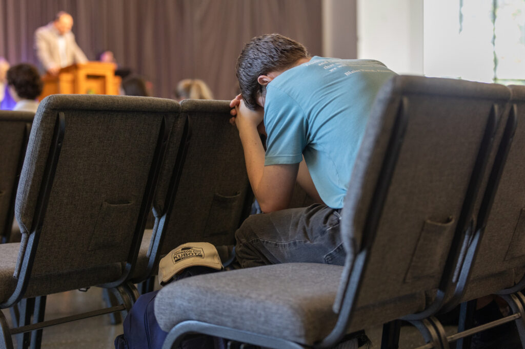 A person in a blue shirt sits in a chair with their head resting on their arms. An event is happening on stage in the background. A hat and backpack are on the chair beside them.