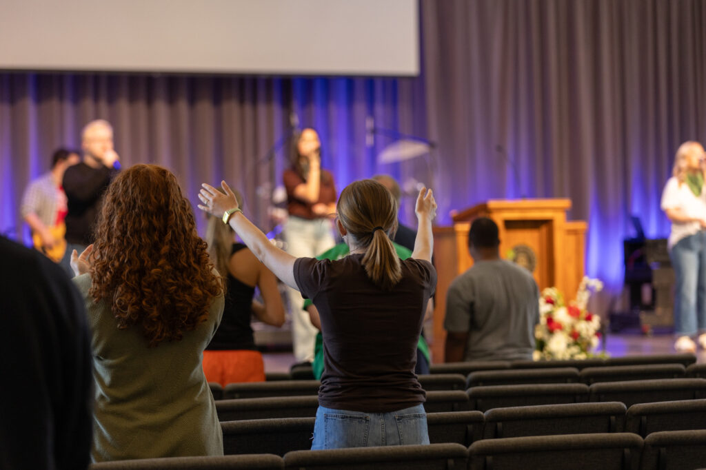 People stand in a church or auditorium, some with arms raised, facing a stage where musicians perform. Rows of chairs are visible, and flowers are arranged near a podium in front of purple curtains.