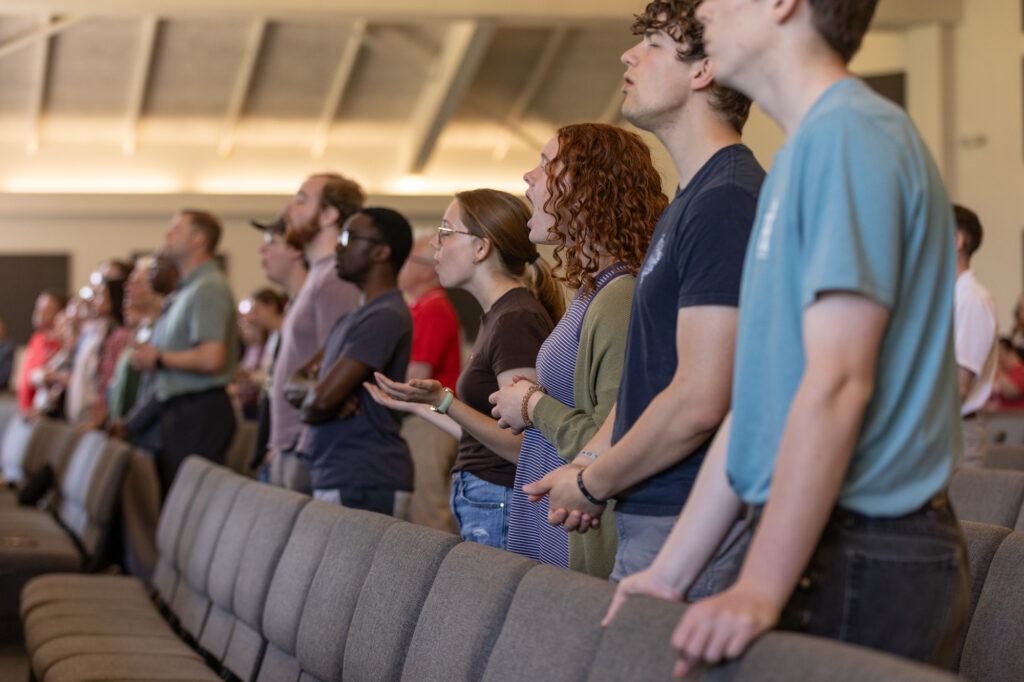 A group of people stands in rows inside a large room, facing forward. Several are singing or speaking, and some hold books or devices. The seats are mostly empty in the foreground. The lighting is bright and natural.