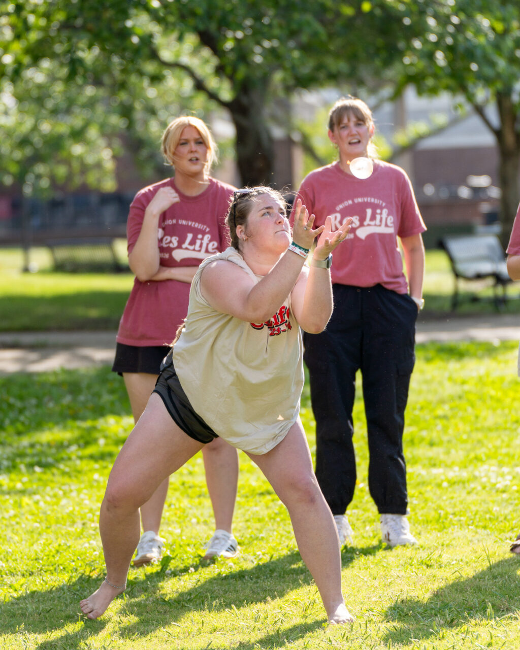 A person in a light tank top and shorts is preparing to catch something outdoors on grass, while two people in matching “Res Life” shirts watch in the background. Trees and benches are visible.