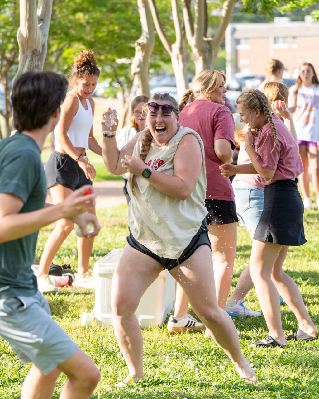 A group of people outdoors on grass having a water fight; one woman in the center is laughing and holding a water bottle mid-splash, while others around her are engaged in playful activity. Trees and buildings are visible in the background.