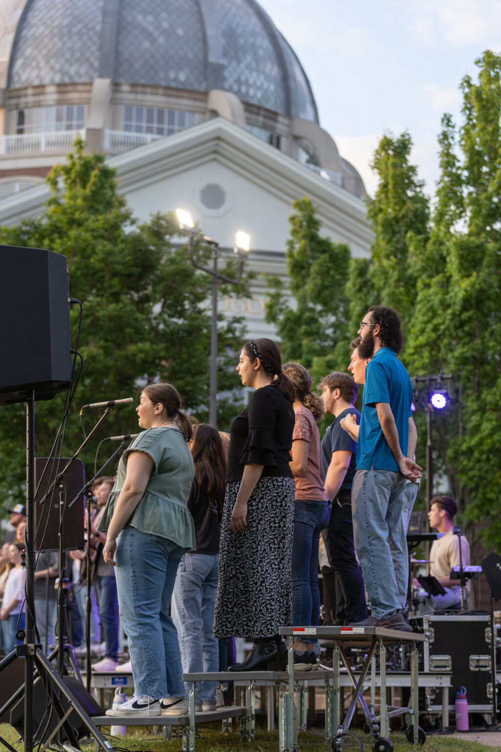 A group of people stand on tiered platforms outdoors, singing into microphones. A domed building and trees are visible in the background. Stage lights and equipment are set up nearby.