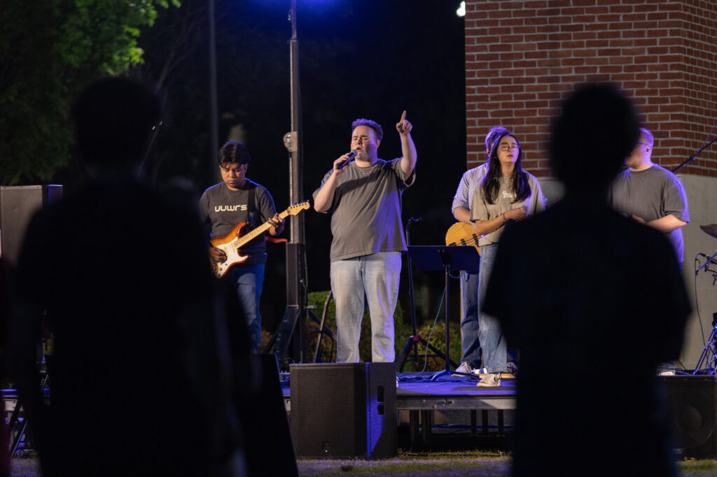 A band performs on an outdoor stage at night. The lead singer holds a microphone and gestures upward. Two other musicians play guitar and bass. Audience members are visible in the foreground as silhouettes.