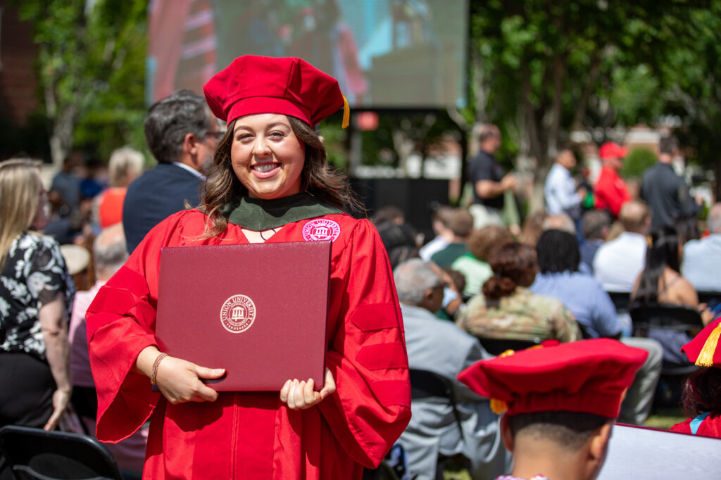 A graduate in a red cap and gown holds a diploma and smiles at an outdoor graduation ceremony—a proud moment made possible by the journey through Graduate Admissions. People are seated in the background, and a large screen displays the event onstage.