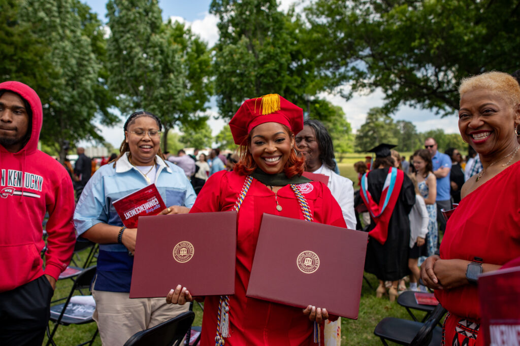 A graduate in a red cap and gown holds two diplomas, smiling proudly, with friends and family standing beside her outdoors—celebrating a milestone made possible through successful Graduate Admissions. Other people and trees are visible in the background at the graduation event.