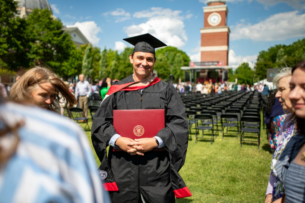A person in a graduation gown and cap holds a diploma, standing outdoors on grass with empty chairs and people around—perhaps moments after celebrating their success in Graduate Admissions. A clock tower is visible in the background under a partly cloudy sky.