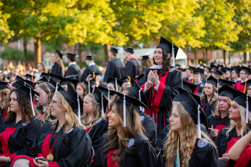 Rows of graduates in black caps and gowns sit outdoors during a graduation ceremony, marking the culmination of their undergraduate admissions journey. One person stands and claps while others sit. Trees and more people are visible in the background.