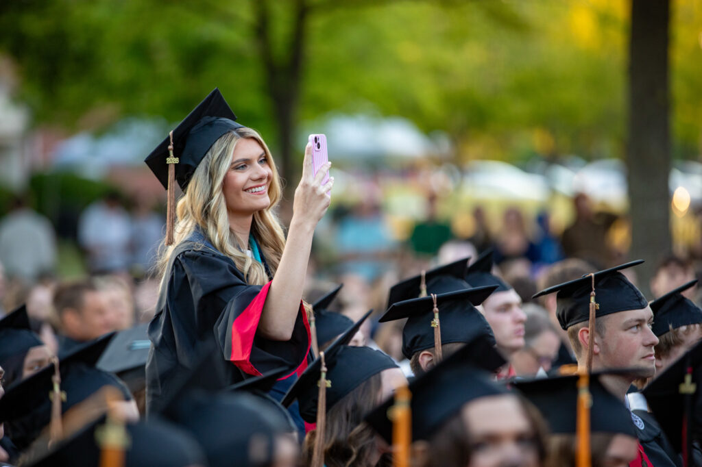 A woman in a graduation cap and gown stands among seated graduates, holding up a pink smartphone to take a photo—a joyous moment celebrating the culmination of her undergraduate admissions journey. The outdoor scene is bright, with green trees and blurred people in the background, radiating the excitement and accomplishment of university admissions success.