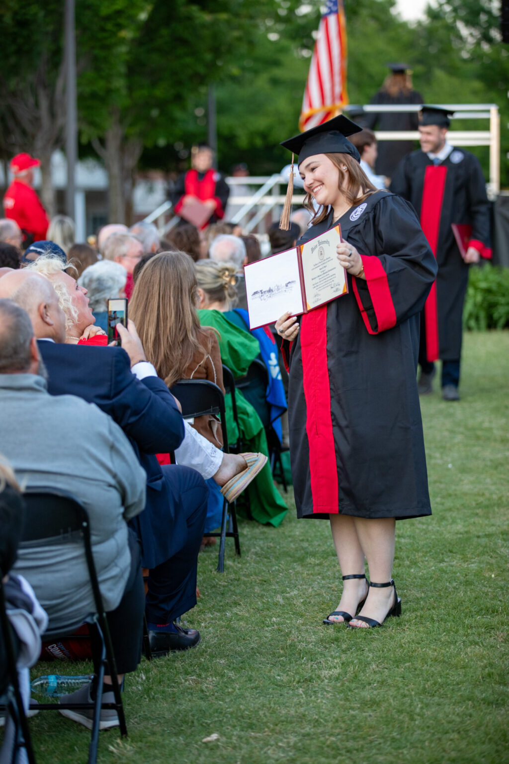 A graduate in a black cap and gown with a red stole stands on grass, smiling and holding a diploma while facing seated audience members at an outdoor graduation ceremony, celebrating the successful culmination of their undergraduate admissions journey.