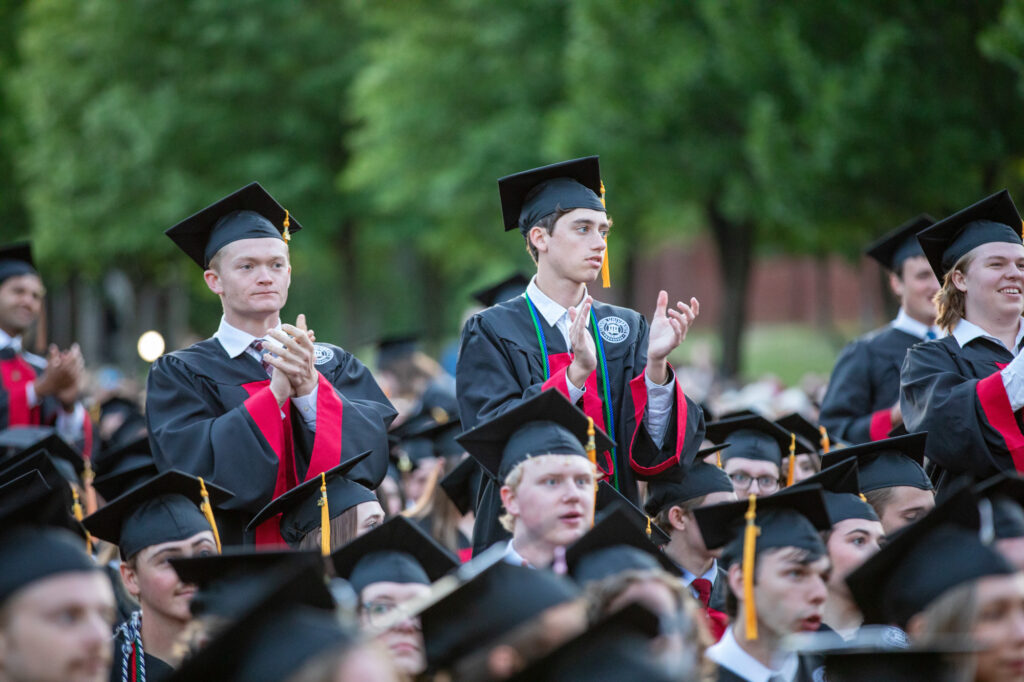 A group of students in caps and gowns clap at an outdoor graduation ceremony, celebrating the culmination of their journey that began with the undergraduate application process. Two students are standing among others who are seated, a testament to their achievements following successful college admissions. Trees and greenery are visible in the background.