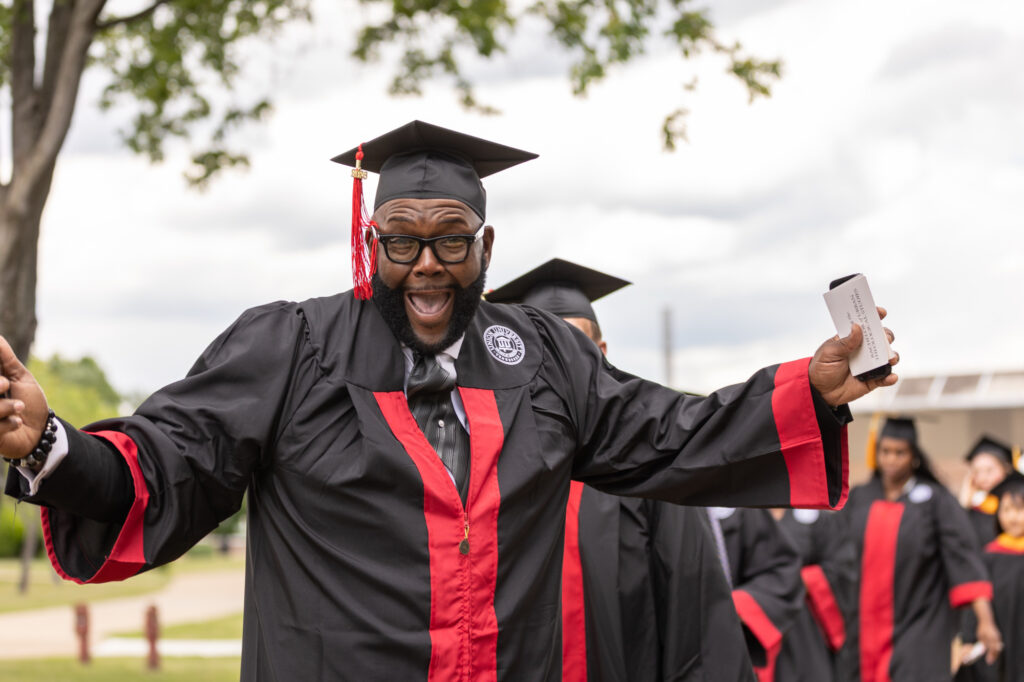 A man in a black and red graduation gown and cap smiles and gestures enthusiastically, holding a phone in one hand, while standing outdoors with other graduates in the background.