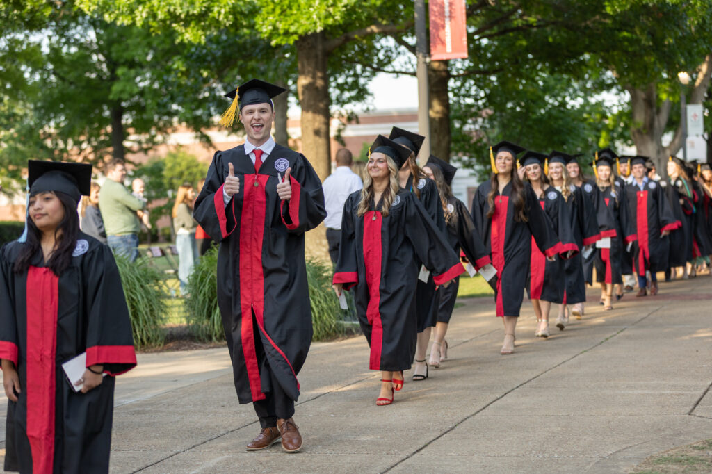 A group of graduates in black and red gowns walk outside on a sidewalk, celebrating their achievements through Undergraduate Admissions. One graduate in front gives two thumbs up and smiles, while trees and people standing in the background add to the festive atmosphere.
