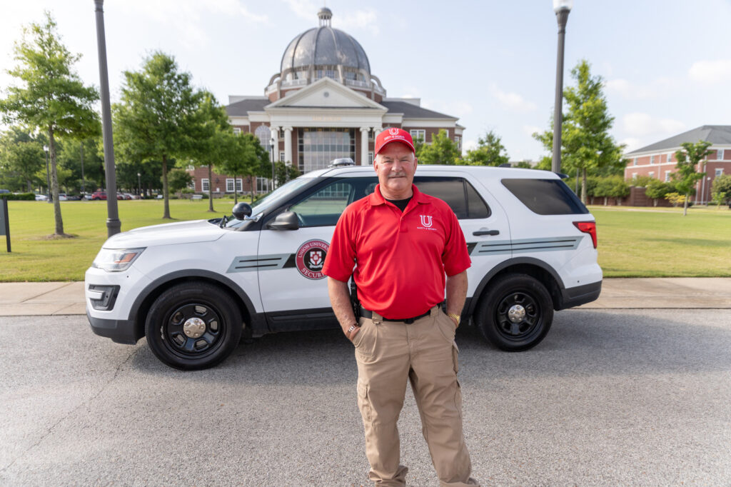 A man in a red polo shirt and cap stands in front of a white SUV with a university logo. A domed building and trees are visible in the background on a sunny day.