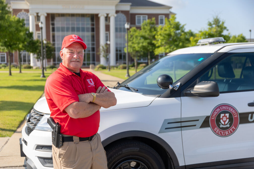 A security officer wearing a red polo shirt and cap stands with arms crossed in front of a Union University security vehicle, with a large campus building in the background.