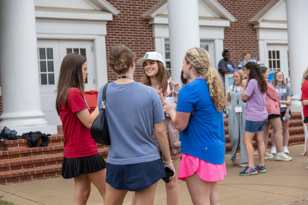 A group of young people stands and talks outside a brick building with white pillars, capturing the spirit of campus living. Some wear name tags and casual clothes, reflecting their involvement in Residence Life. Others gather and chat in the background, enjoying the vibrant atmosphere of student housing.