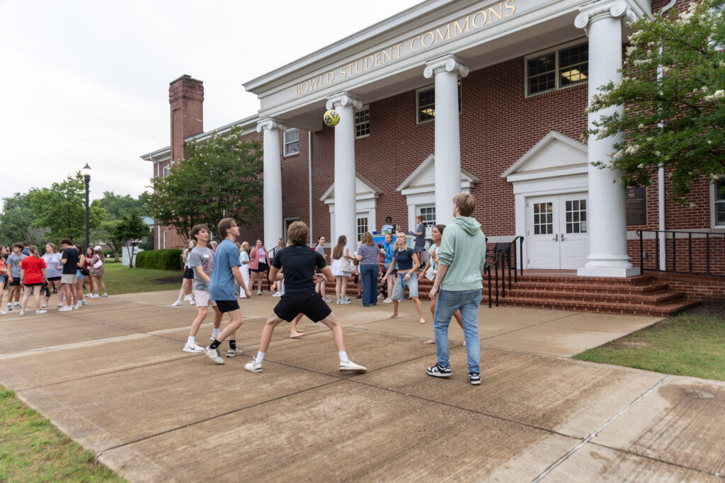 A group of people involved in Residence Life play with a soccer ball outside a building labeled Powell Student Commons. Others are gathered near the entrance, with trees and grass visible nearby. The weather appears overcast.